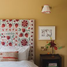 An earthy yellow-painted bedroom with a wall light above the bedside table and a floral-print fabric headboard