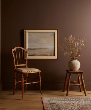 dark brown wall in living space with wooden chair, wooden stool with vase of dried stems and landscape framed picture on wall