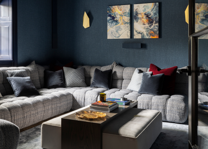 A living room corner with dark blue walls and an l-shaped gray sofa with a gray ottoman in the center with a table over the top across the middle styled with books and decorative objects