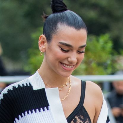 Dua Lipa has her hair in a high bun and wears an off-the-shoulder striped top with a lace bra underneath