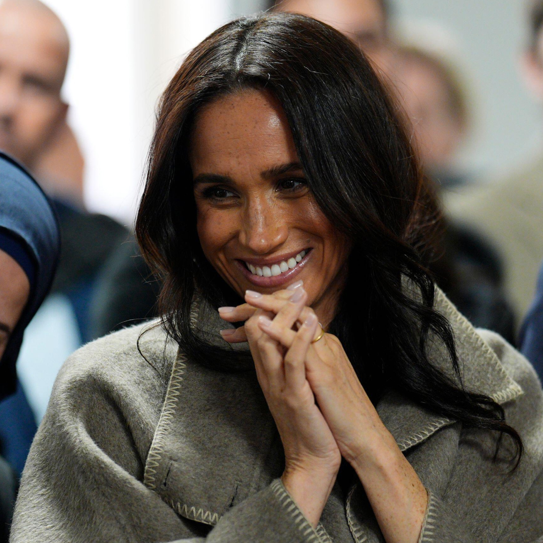 Meghan Markle clasping her hands and smiling in a taupe coat