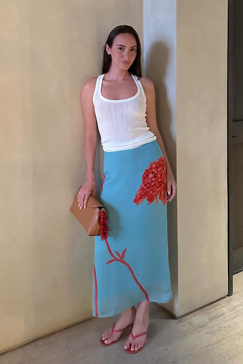 a woman wearing a white tank top, blue and red skirt, red flip-flops, and a tan clutch bag