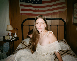 ethel cain wears a white vintage dress sitting on a bed next to an american flag in a promo shot for preachers daughter