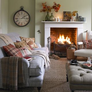 Living room with light green walls and a fire burning in the fireplace which has a white mantle, and cosy sofas and armchairs with cushions in front, and a circular roman numerals clock on the wall