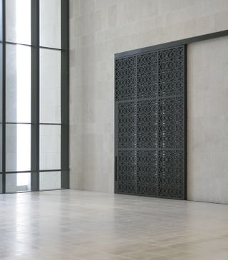 A tall black patterned door on grey concrete wall. on the left of the wall is a clear floor to ceiling window with black metal panel detail