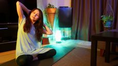 Woman sits cross-legged on exercise mat, with her hands behind her head, eyes closed and torso tipped to one side. The curtains are drawn and a corner light casts a low light