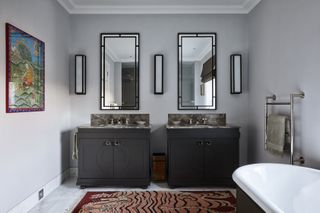 Bathroom with freestanding bath, twin vanities in gray with mirrors and lights over, rug on floor, artwork on wall and neutral walls and flooring