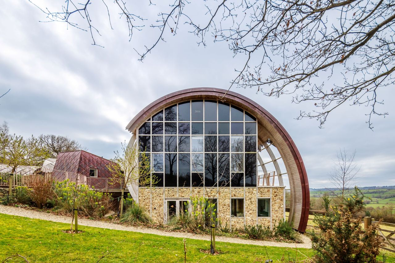The front of the house shows a stone wall with large window above it with a balcony on the side with a Fibonacci, curved shape