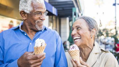 An older couple laugh together as they walk along a city street and eat ice cream.