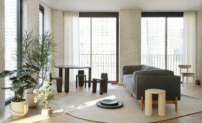 A photo of an apartment lounge with tall windows and furniture, including: a black round three-legged table with small-back chairs; a grey sofa, a collection of indoor plants and a yellow side table. 