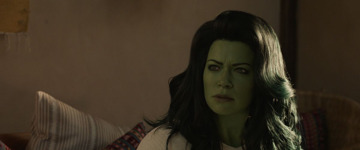 Tatiana Maslany as She-Hulk, sitting on a sofa in Hulk mode and staring at someone off-camera to her right.