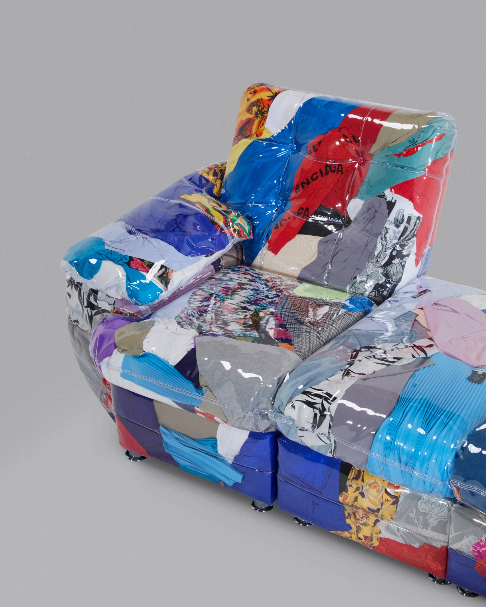 A transparent-vinyl couch filled with disused colorful clothes, against a gray background.