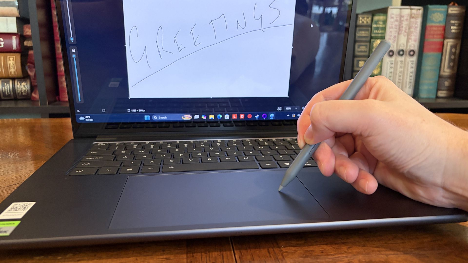 A hand writes “GREETINGS” on a Lenovo Pro 9n touchpad with a stylus, surrounded by bookshelves; the casual scene feels focused and creative.