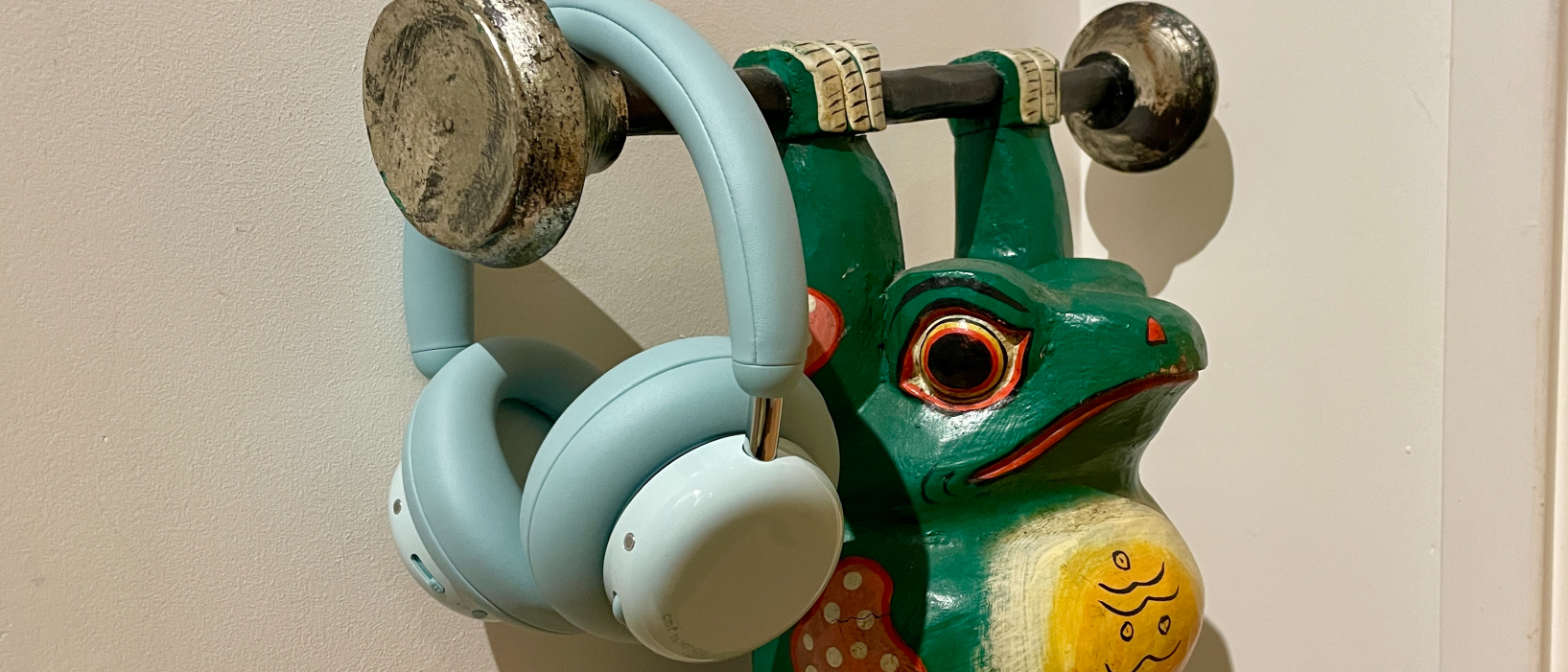 CMF Headphone Pro in pistachio green held in a hand or hung on a barbell, being pushed up by a carved frog 