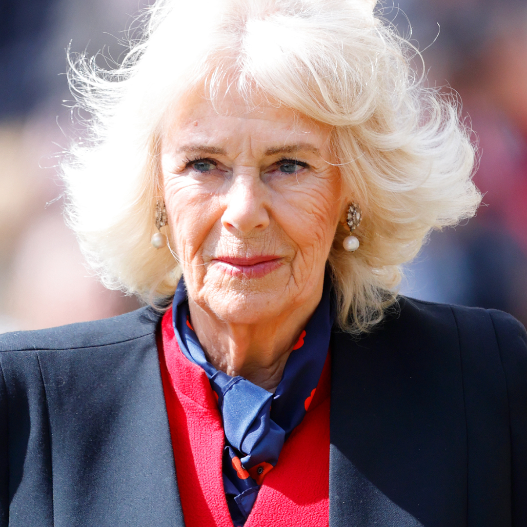 Queen Camilla wears a black blazer and a red dress and has blonde hair which is in an upheaval in the wind