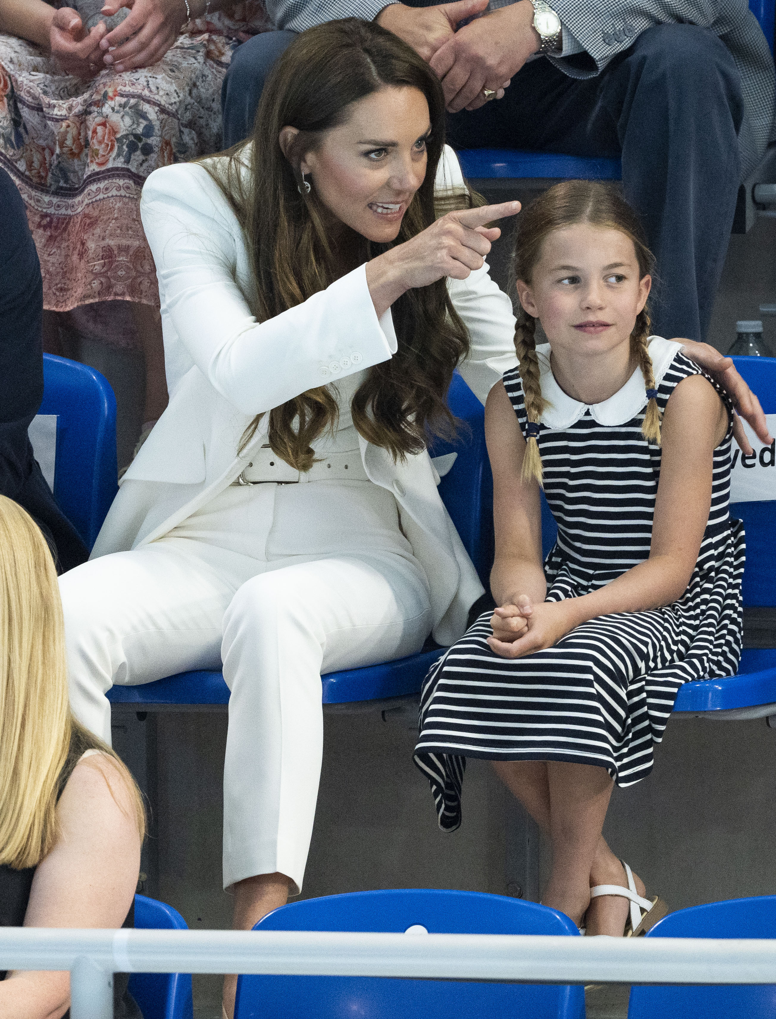 Princess Charlotte wearing a striped dress sitting next to Princess Kate in a white suit