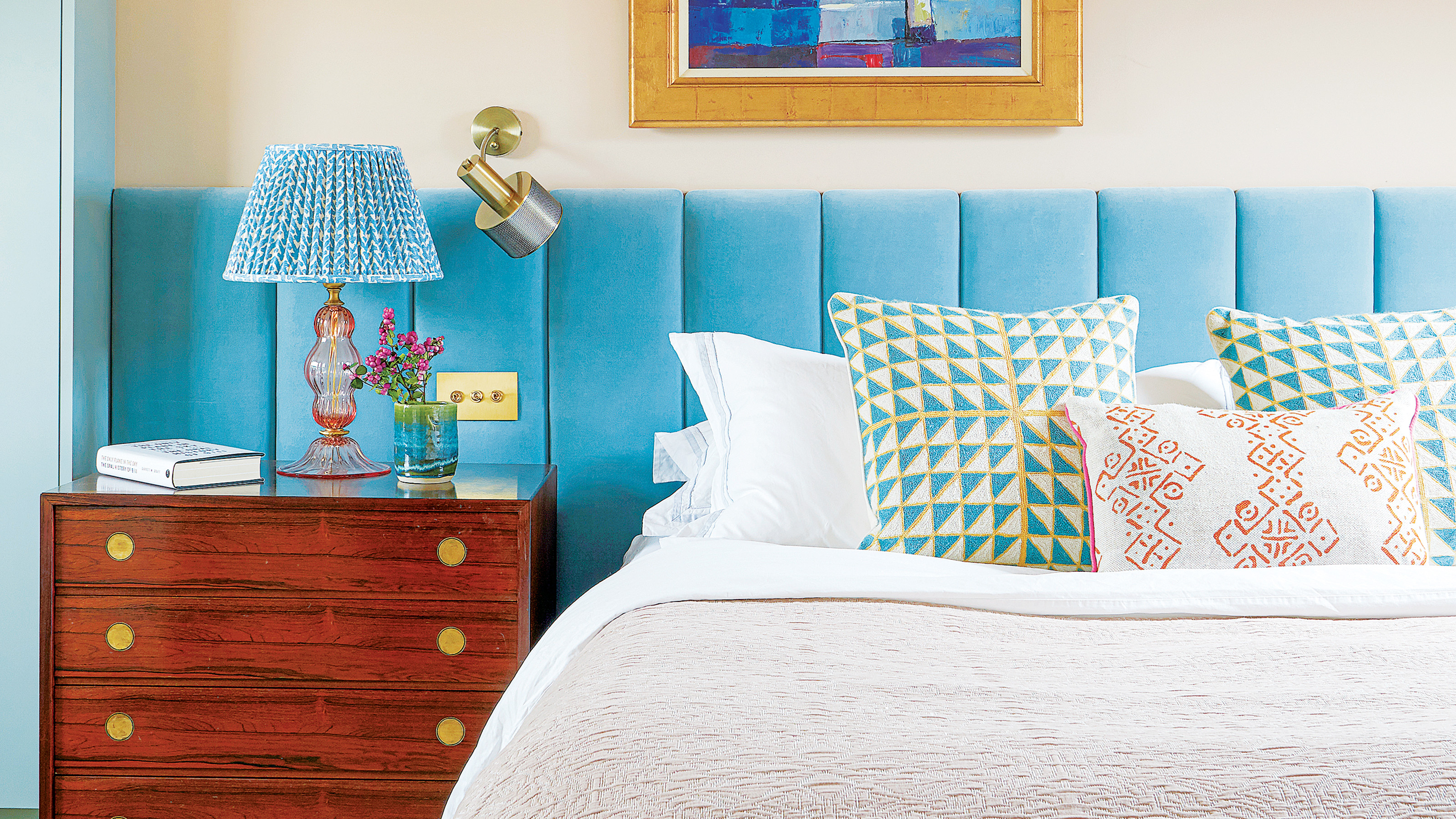 bedroom with cream walls and abstract artwork above cream and whtie bed and an oversized wide headboard in a sky blue velvet reaches the full width of the wall and behind a wooden bedside table 