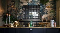 Moody dark kitchen with gleaming gold counters and glass-fronted wall cabinet, against a dramatic wallpaper with tree scene