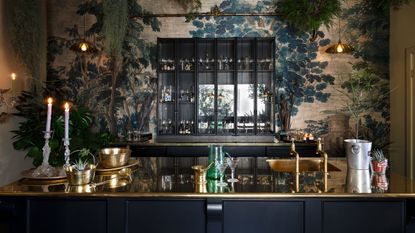 Moody dark kitchen with gleaming gold counters and glass-fronted wall cabinet, against a dramatic wallpaper with tree scene