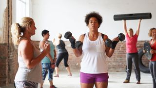 Woman lifting dumbbells into a curl with the help of an instructor during a workout class, representing strength training for weight loss