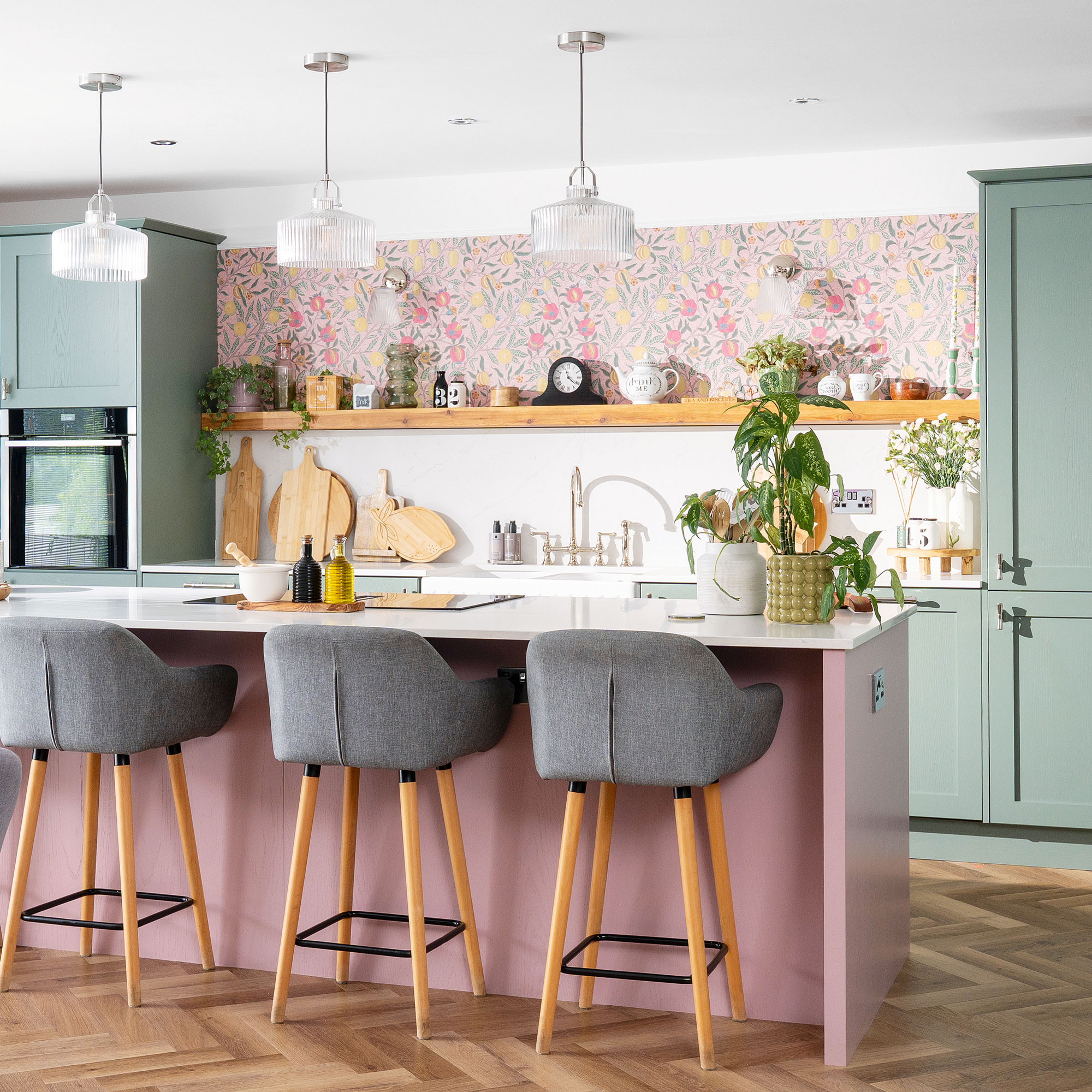 Mint green kitchen cabinets next to pink floral wallpaper, both are behind a pink kitchen island with white countertop and grey stools