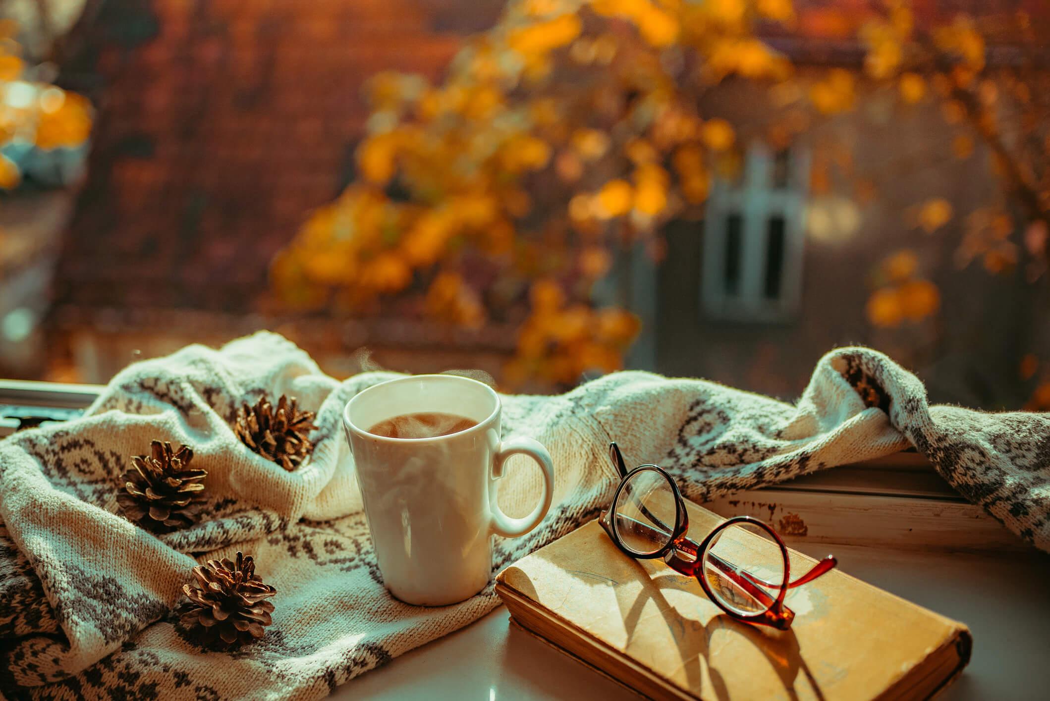  A cosy autumn scene by a window with a book, a pair of glasses, and a warm drink on a blog article on autumn activities.  