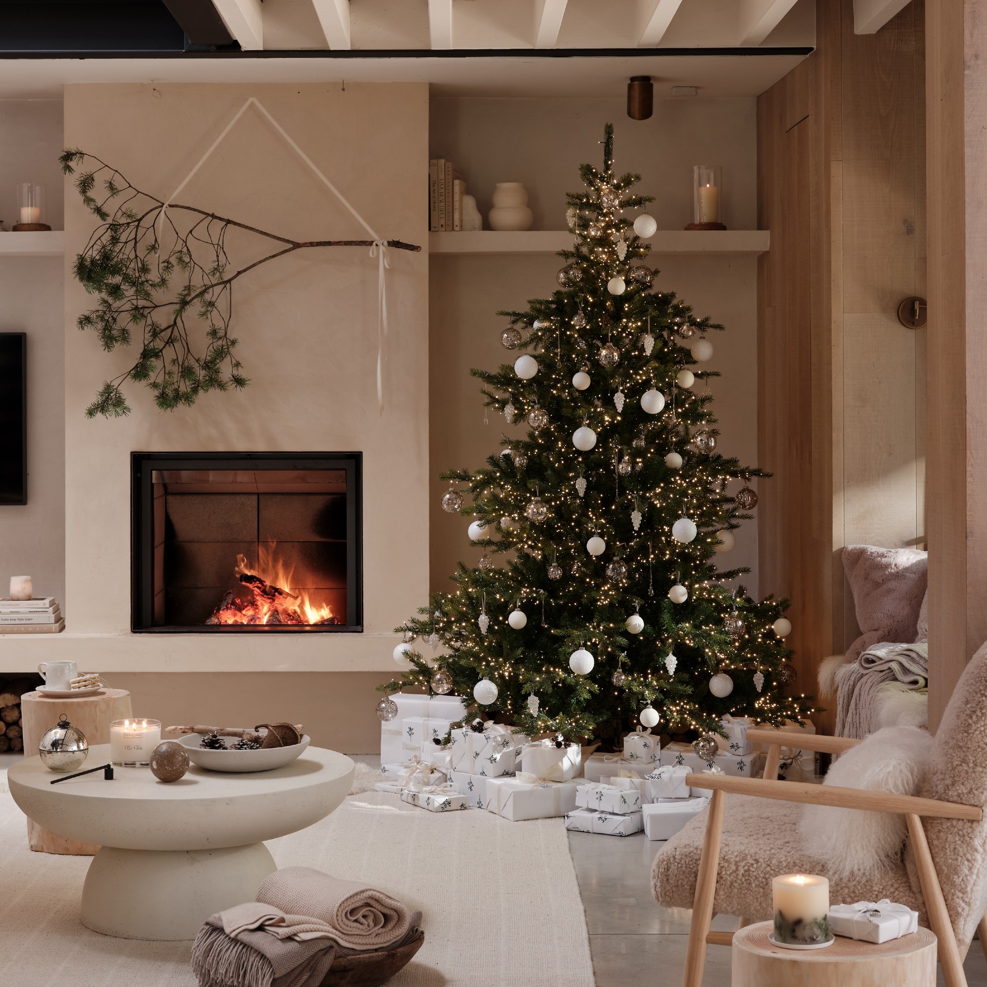 The White Company's extra bright Christmas tree in a modern living room with a lit fireplace and a decorative branch hung above it