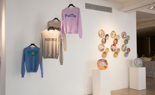 Beige, blue and pink jumpers on hangers hanging from a wall next to a wall display of colourful plates