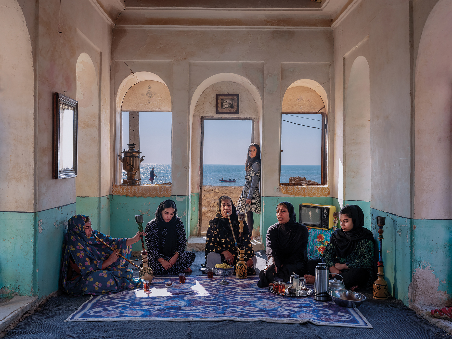 A group of women in traditional attire sits in a sunlit, sea-view room, enjoying tea and conversation, with vintage decor surrounding them