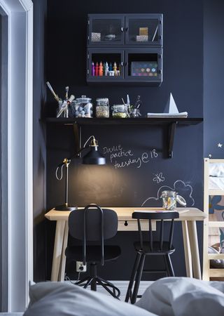 A small desk area in an alcove with dark walls, with floating shelves full of craft essentials, and a cupboard above with art supplies in it