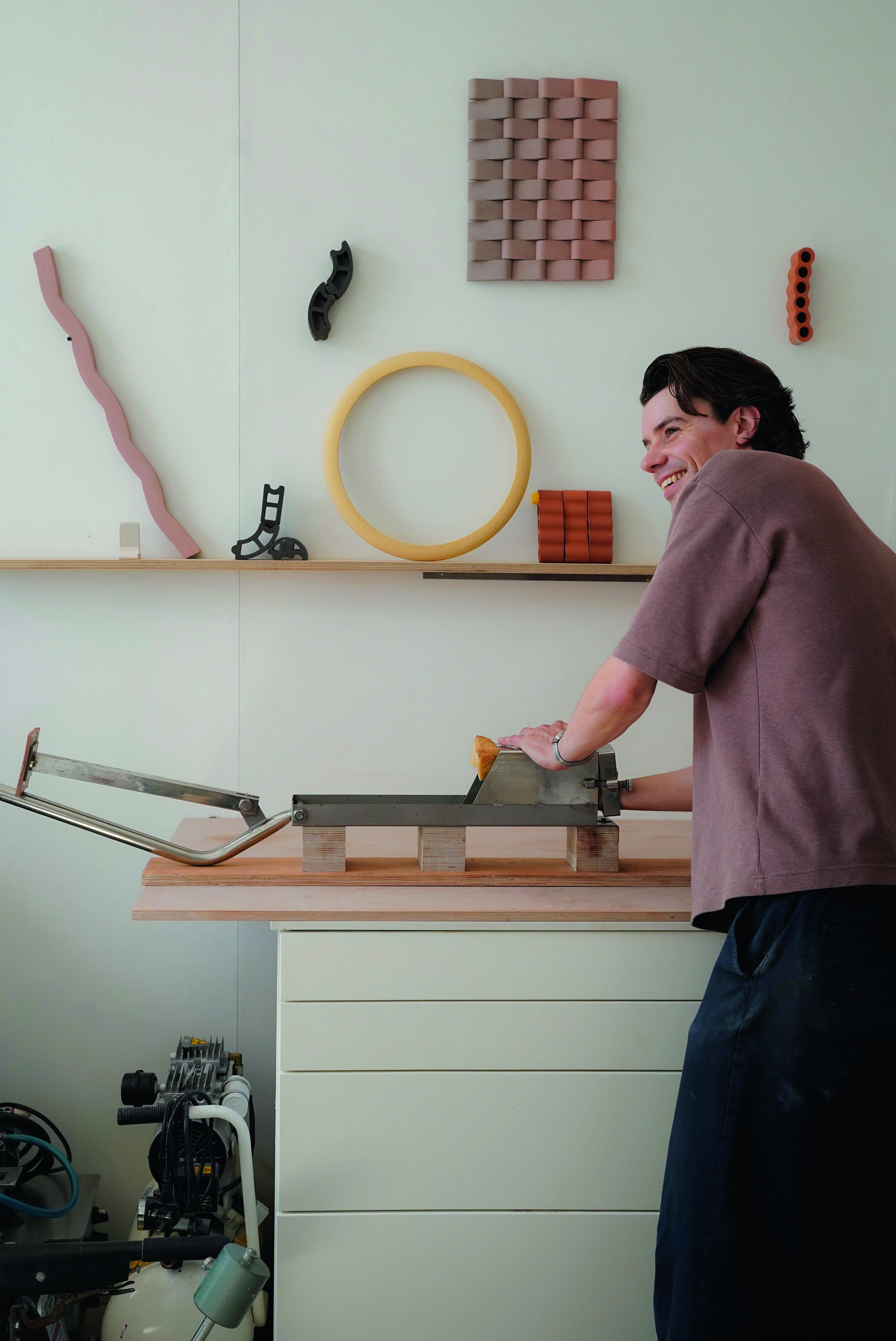 A man works in a wood workshop dressed in loose workwear in brown and navy blue while smiling in front of a wooden shelf.