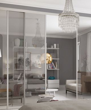 transparent room dividers separating a living room into two separate spaces, one with a white display unit and gold lamp and the other showing the reflection of a bed and crystal chandelier