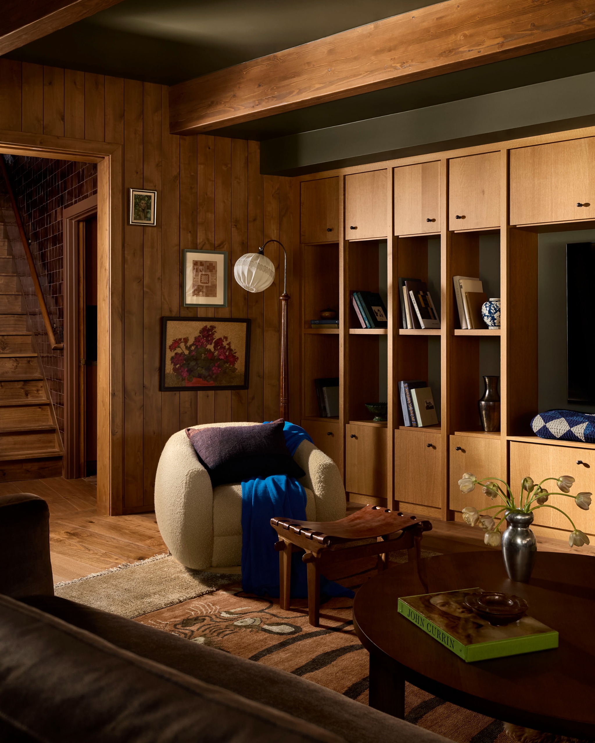 A cozy living room clad in wood with a cream boucle tub accent chair, built-in storage and shelving and a large patterned rug, with framed artwork on the wall behind the chair