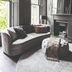 A grey living room with a curved grey velvet sofa and a matching pintucked ottoman on a round grey rug