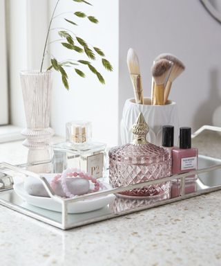 A silver bathroom tray with a soap tray, a pink storage glass jar, a square perfume bottle, and a white ceramic pot with makeup brushes in it