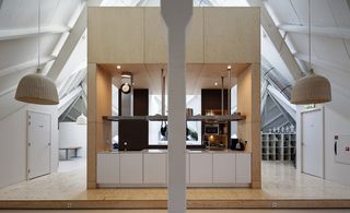 Alternative interior view of the kitchen area housed in a light and dark coloured unit at the Super Cube. The kitchen features white cupboards, a suspended metal shelf with pots and pans, appliances and spotlights. Outside the kitchen area are pendant lights and a white pillar