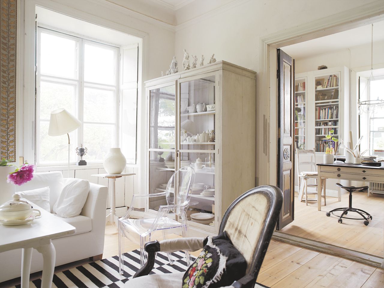 living room apartment with view into office, white couch, display cabinet, armchairs, rug, white accessories, pale wooden floor 