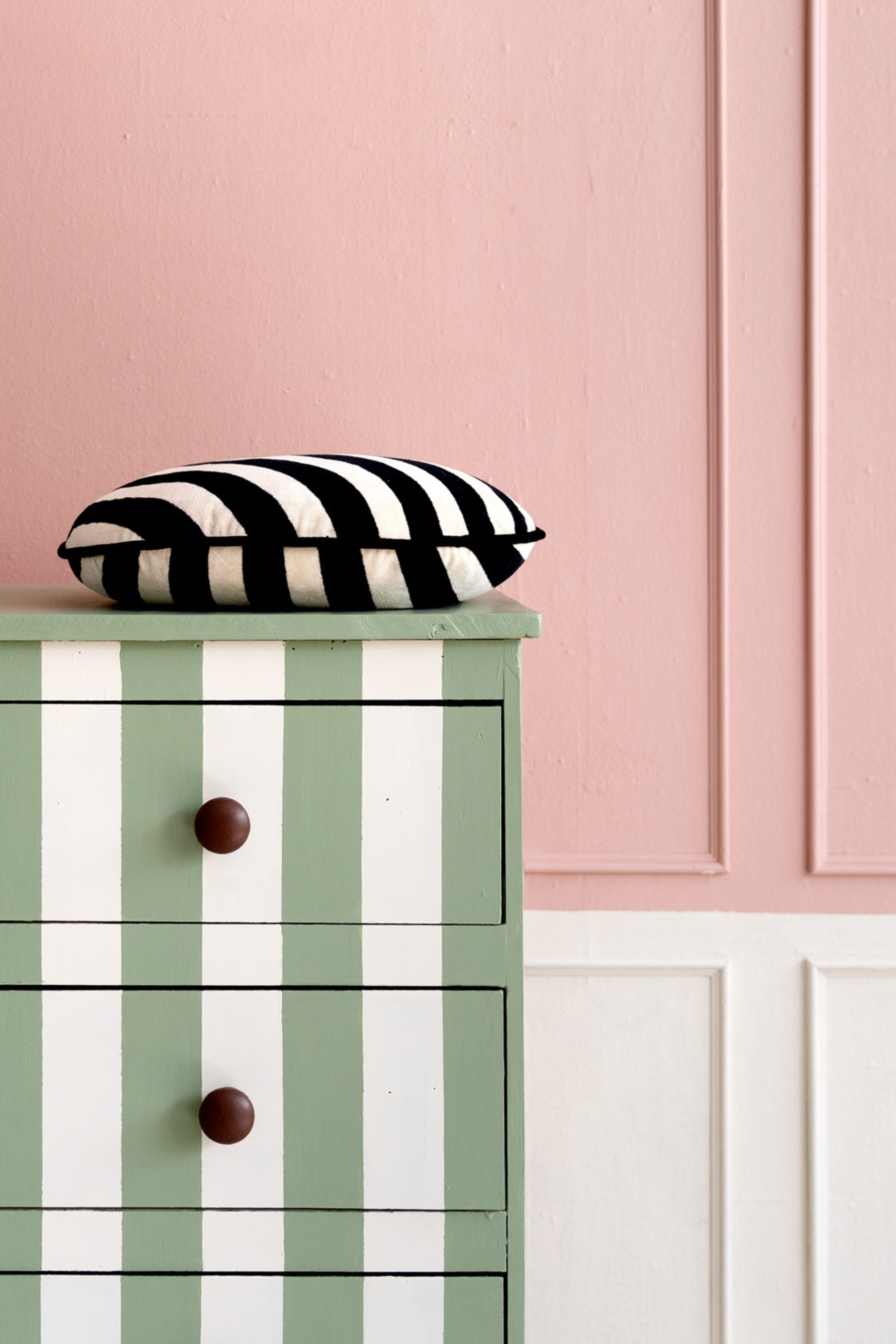 Close-up image of a dresser painted in green and white stripes with dark wooden knobs. There is a black and white striped pillow on top of the dresser. The walls are pink and white color block.