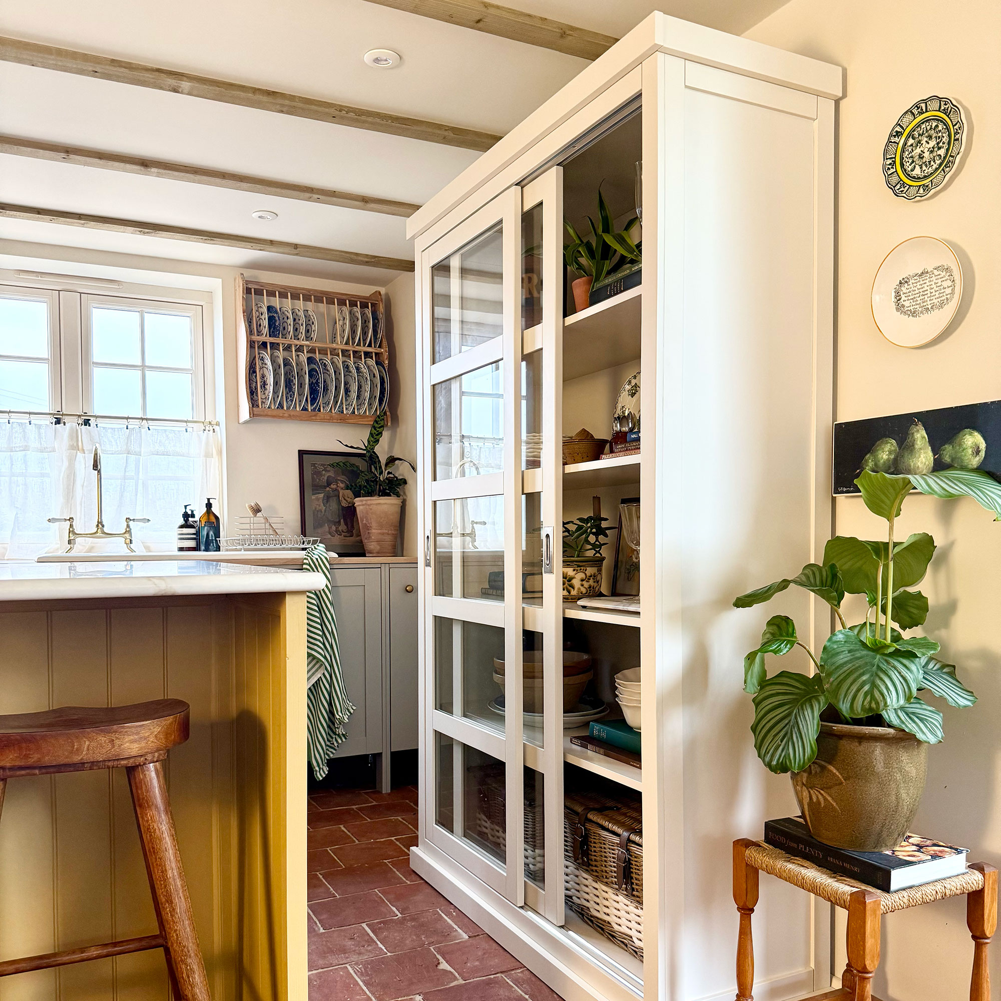 Kitchen with white, glass fronted cabinet, wooden plate rack on walls, terracotta floor and island with yellow panelling