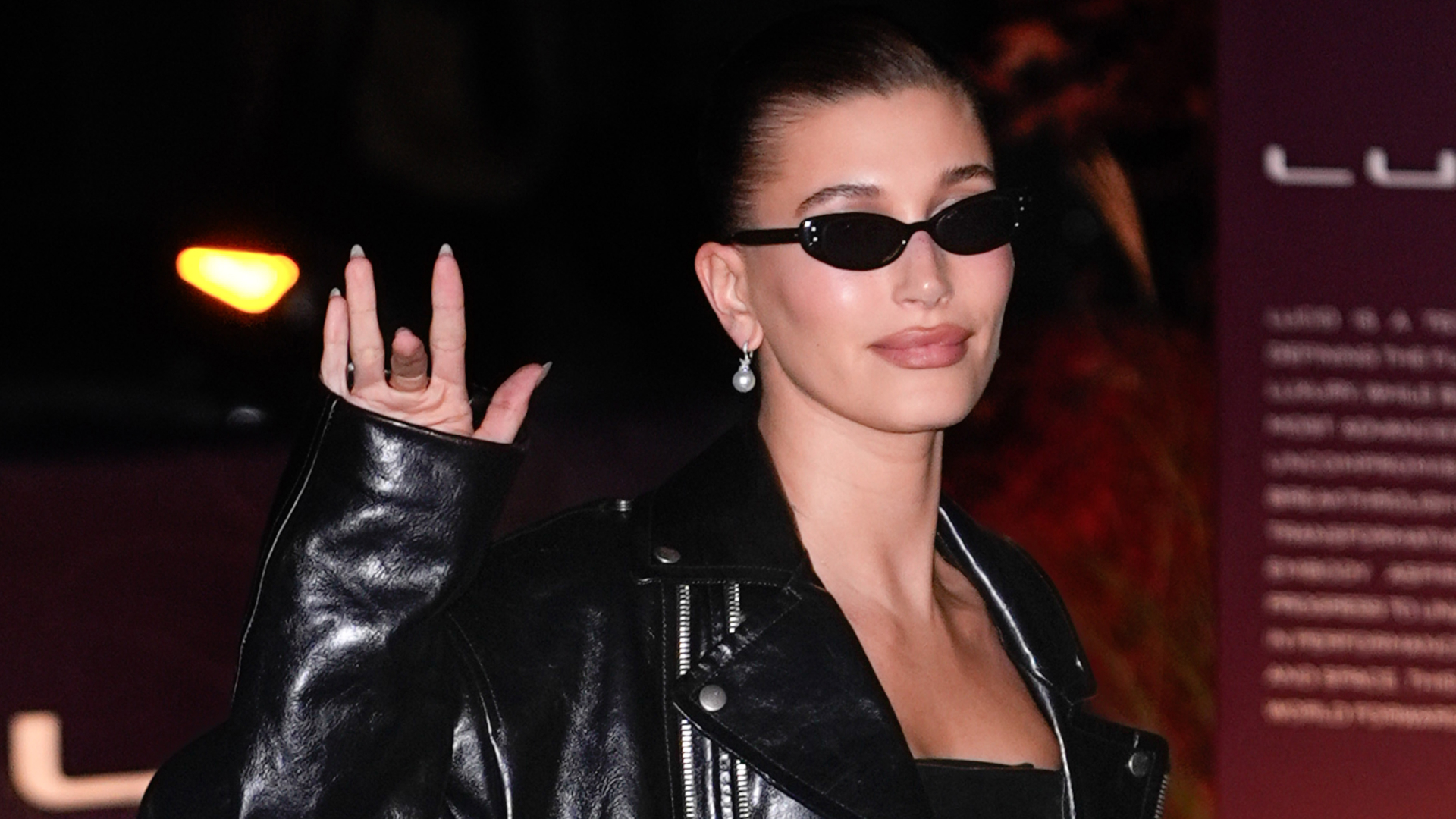 Hailey Bieber wearing a leather jacket and black sunglasses. She is waving her fingers at the camera