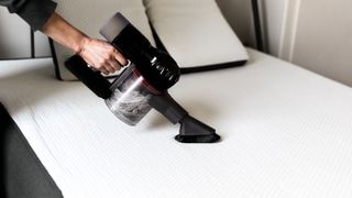 A hand holding a small portable vacuum, and vacuuming the surface of a mattress