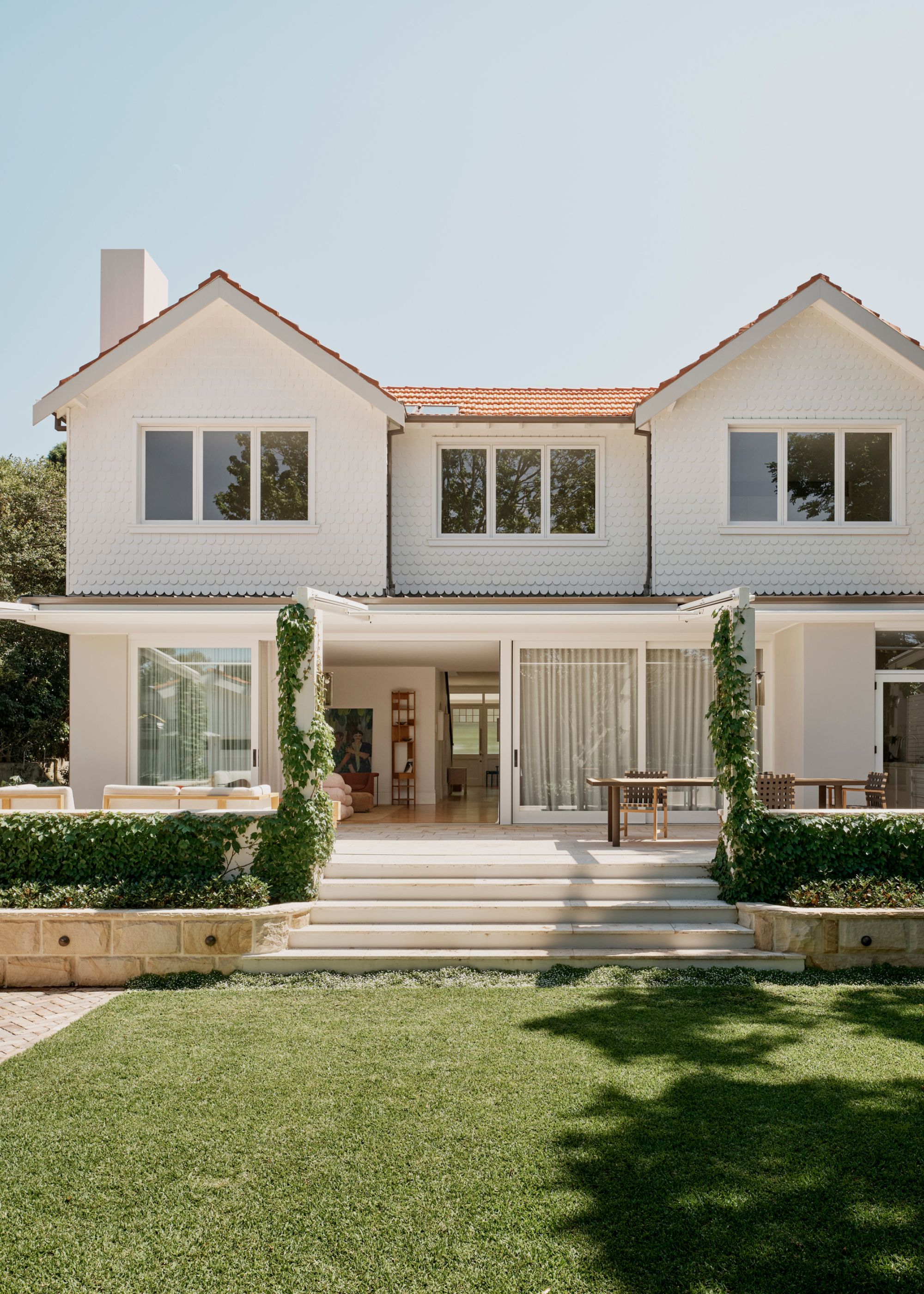 A large two-story home with a white facade, and a spacious garden with trailing plants and a green lawn