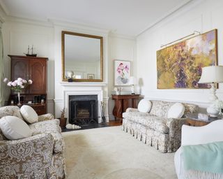 A living room with white walls, patterned sofas, art on the walls, traditional wooden cabinet and marble fireplace