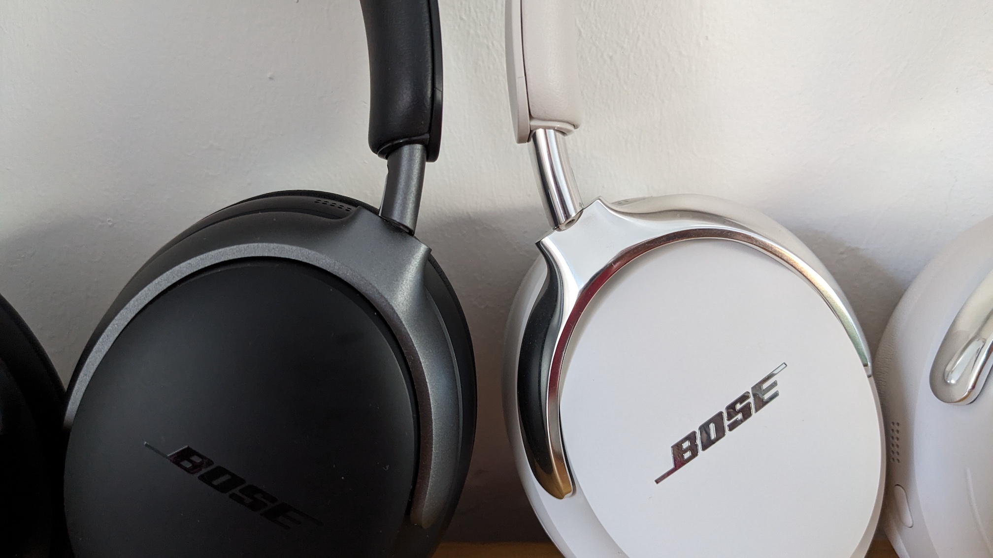 A close up of a white pair of Bose QC Ultra Headphones (2nd Gen) standing next to their black 1st Gen predecessors in front of a white wall.