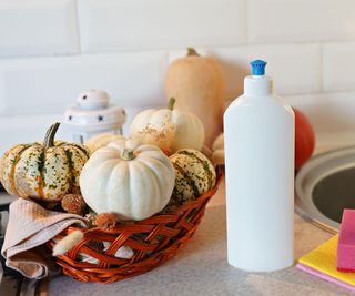 A basket of pumpkins beside a bottle of soap and sponges in a kitchen