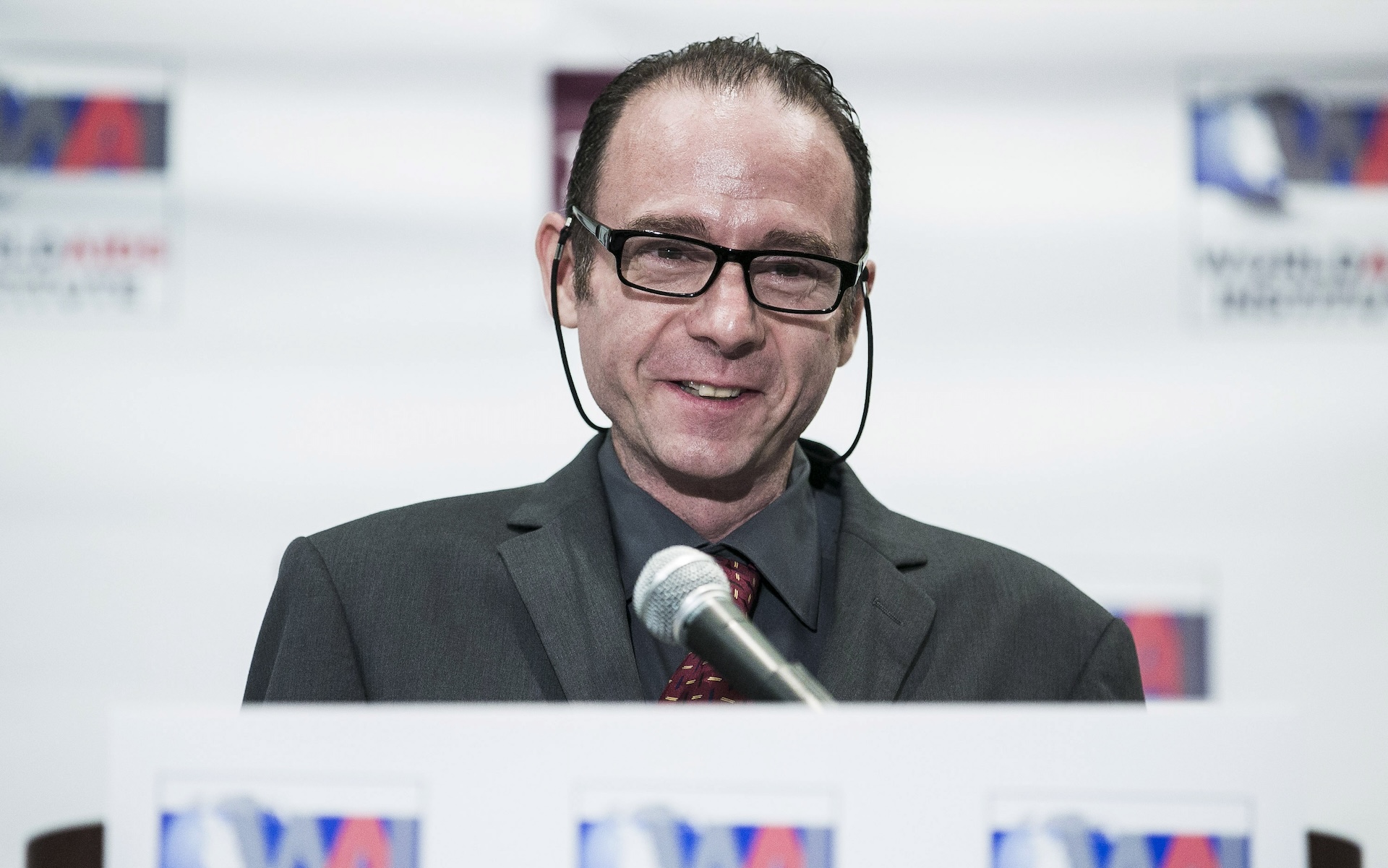 A photo of a smiling man wearing glasses and a suit and standing at a podium to speak