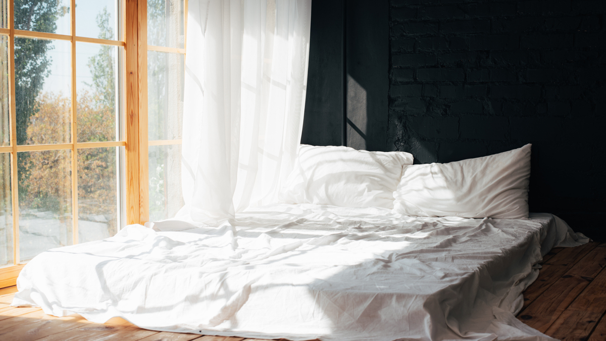 A mattress with white bedding on the floor next to a window with lots of natural light