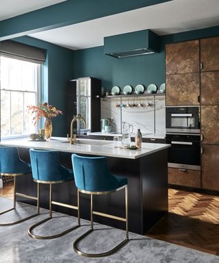 An example of how to plan a layout for a small kitchen showing a kitchen with dark green walls and cabinets, an island with gold and blue bar stools and bronze kitchen cabinets