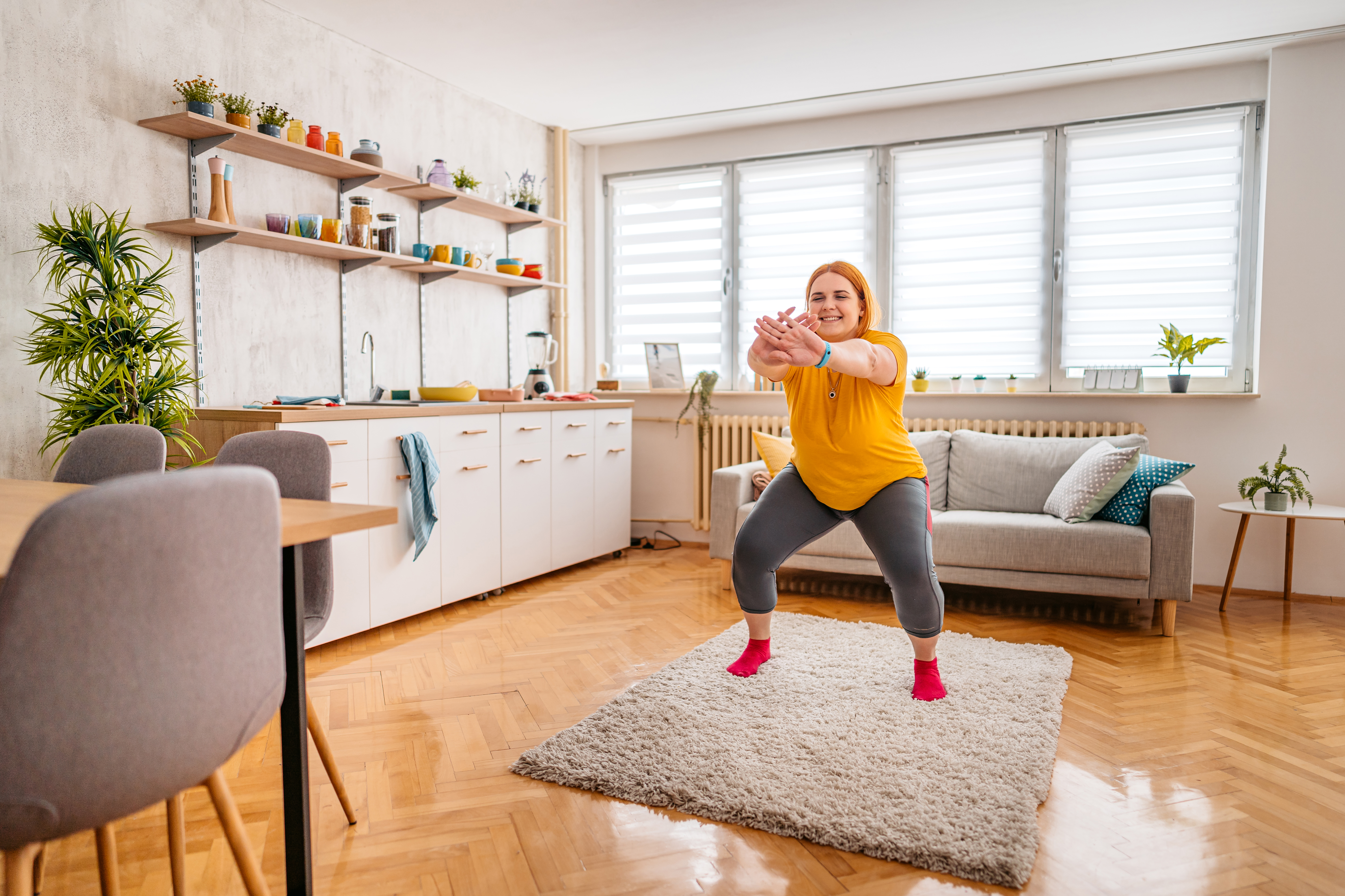 A woman wearing leggings and a t-shirt practices squats at home. She is standing on a rug, behind her we see a couch and to her side there is a kitchen unit with a sink and dishes. 