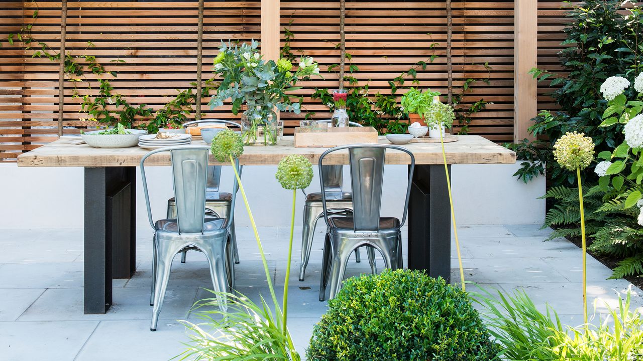 Garden dining table with metal bistro chairs on a light paved patio with wooden fence panelling behind